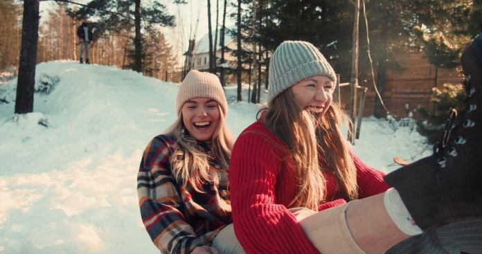 Winter holiday fun. Two beautiful excited happy women smile sledging on snow forest slope towards camera slow motion.